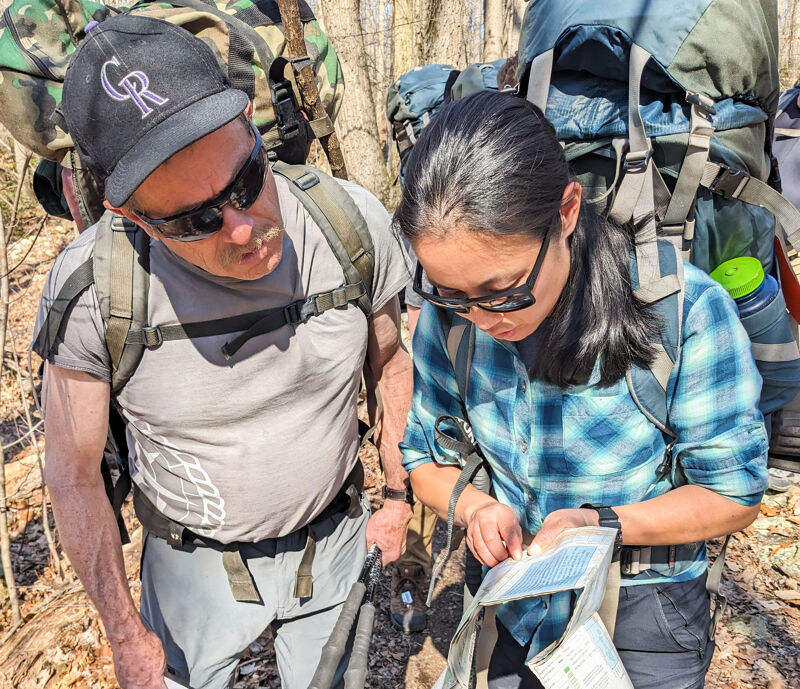 Two hikers are looking at a map in a forest. The man on the left is wearing a baseball cap, sunglasses, and a gray t-shirt. The woman on the right is wearing a blue plaid shirt and glasses. Both are wearing backpacks, suggesting they are on a multi-day hike. They appear to be consulting the map to navigate their route.